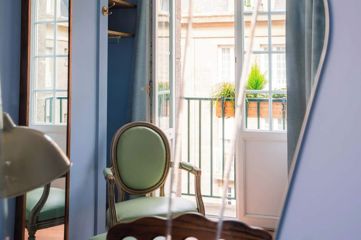 A room with a blue wall, a green chair, a wooden table, and a window with a view of buildings and plants outside.