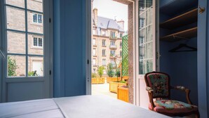 A room with a bed, a chair, and a view of an outdoor balcony with wooden furniture and a building in the background.