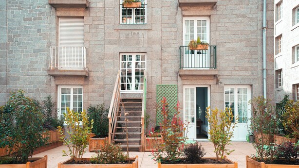 A multi-story building with a brick facade, wooden balconies with hanging planters, and a central staircase leading to the entrance.