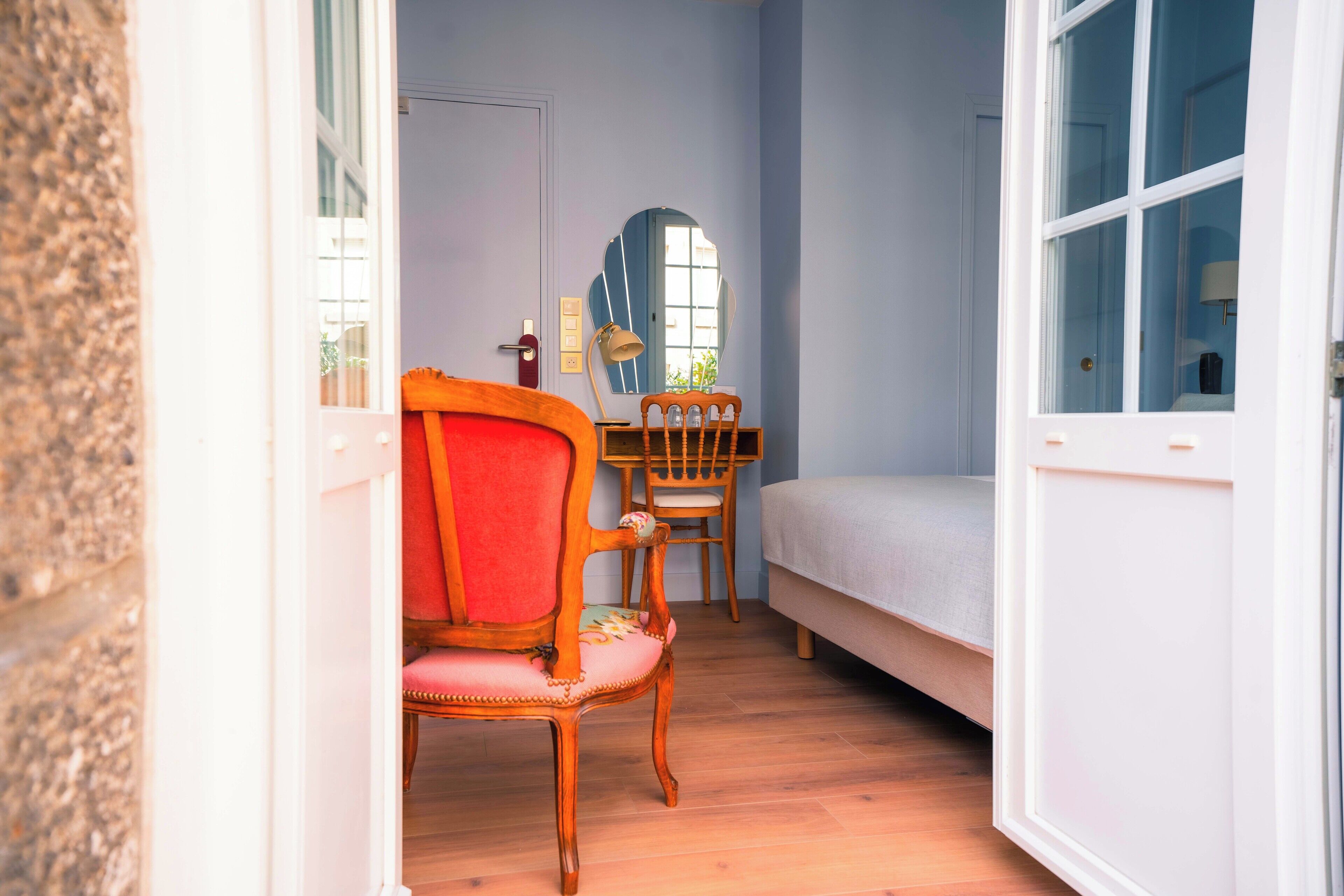 A bedroom with a bed, a wooden chair, a desk, and a mirror.