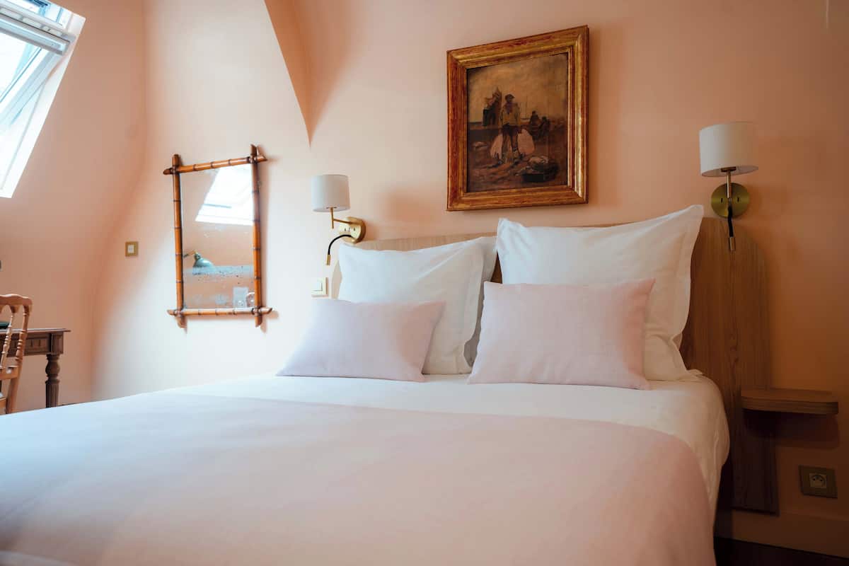 A neatly made bed with white linens, a painting above it, and a lamp on the nightstand.