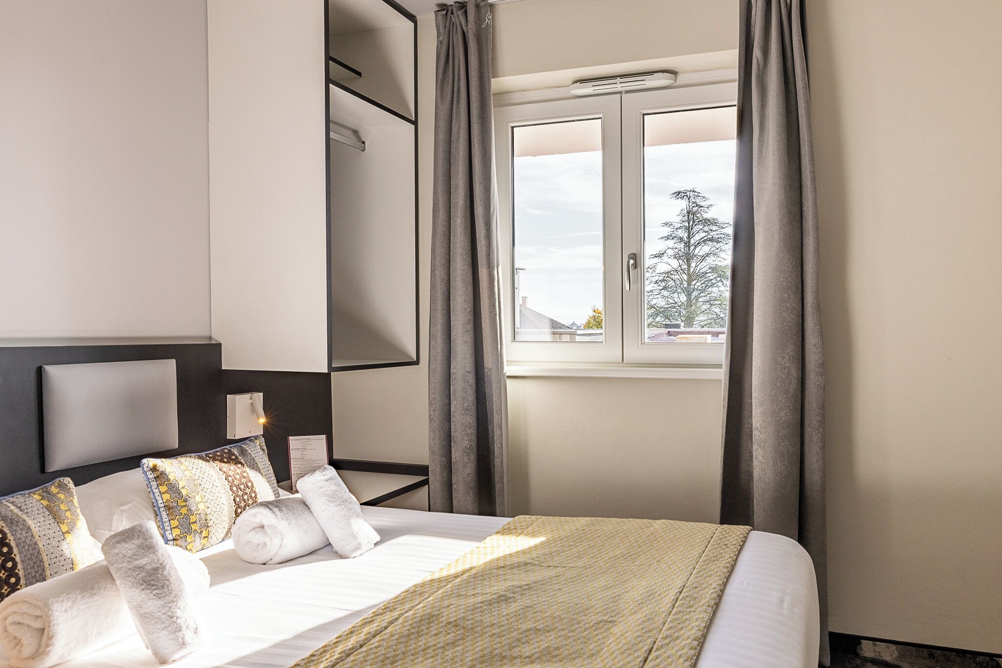 A hotel room with a bed, a window with curtains, a wall-mounted air conditioner, and a view of trees outside.