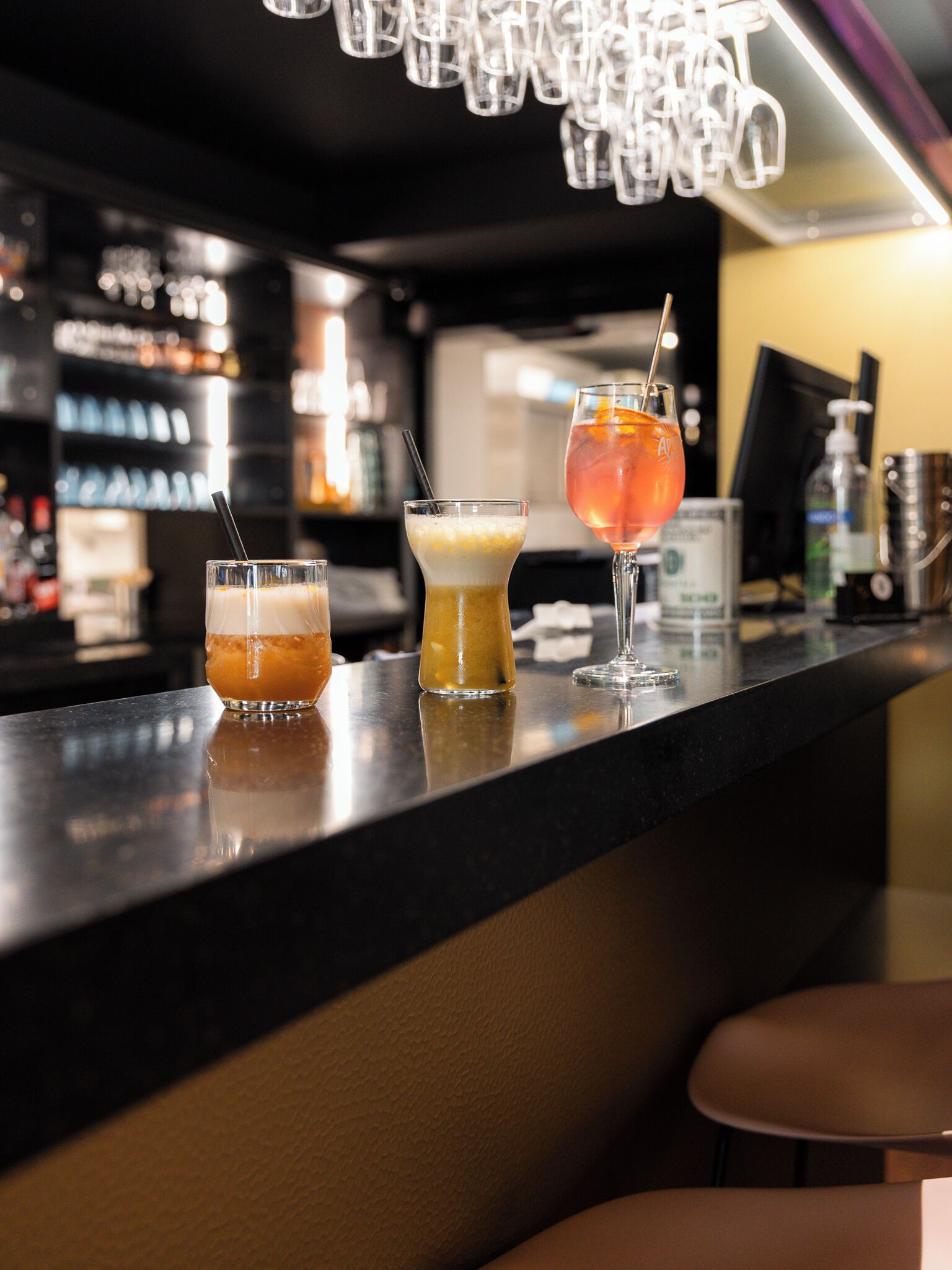 A bar counter with three drinks, a bottle of hand sanitizer, and a cash register.
