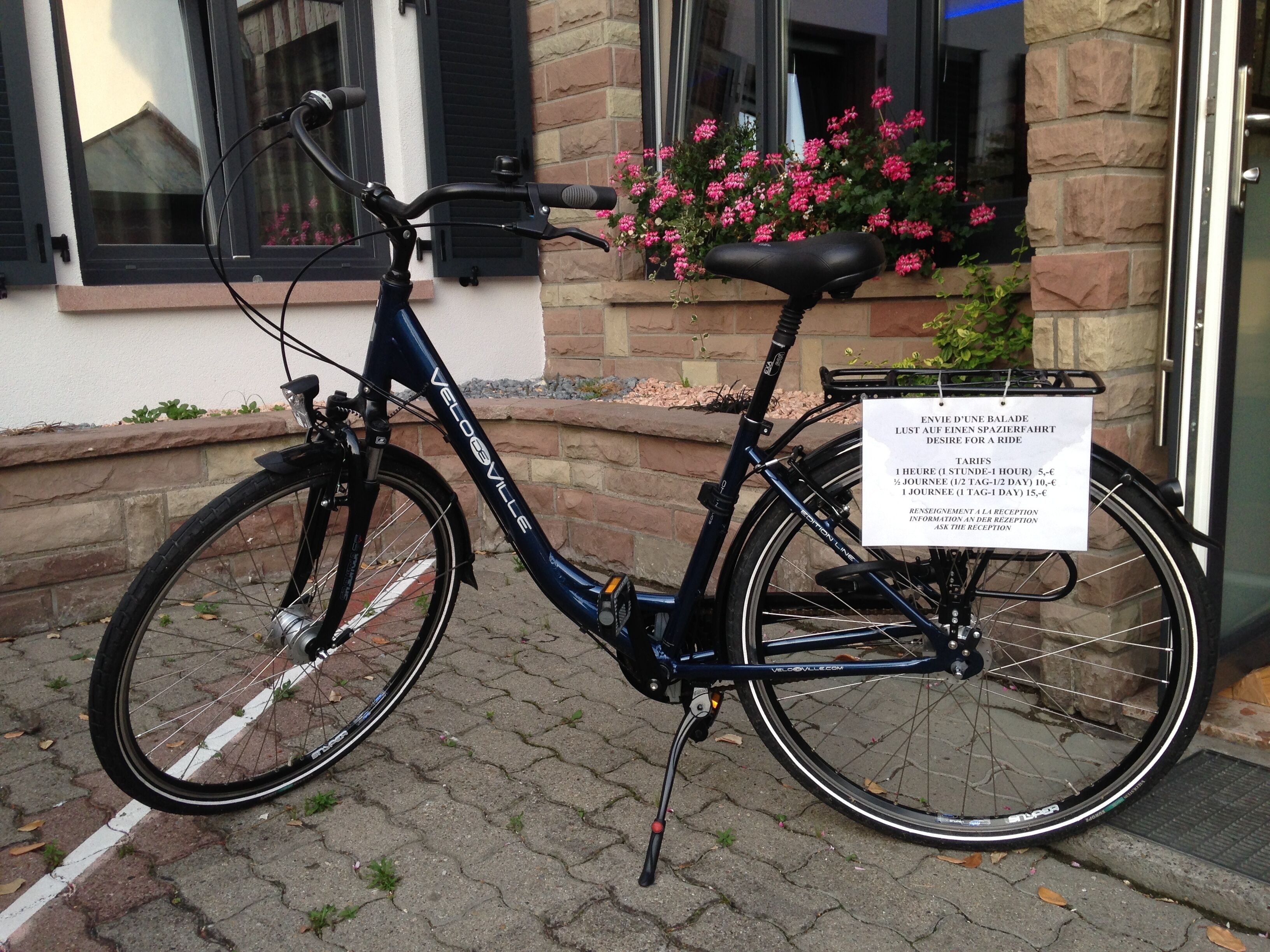 A bicycle with a basket and a sign attached to the back, parked on a cobblestone path in front of a building with a window and flower box.