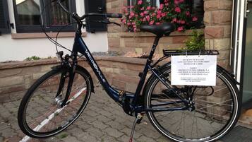 A bicycle with a basket and a sign attached to the back, parked on a cobblestone path in front of a building with a window and flower box.