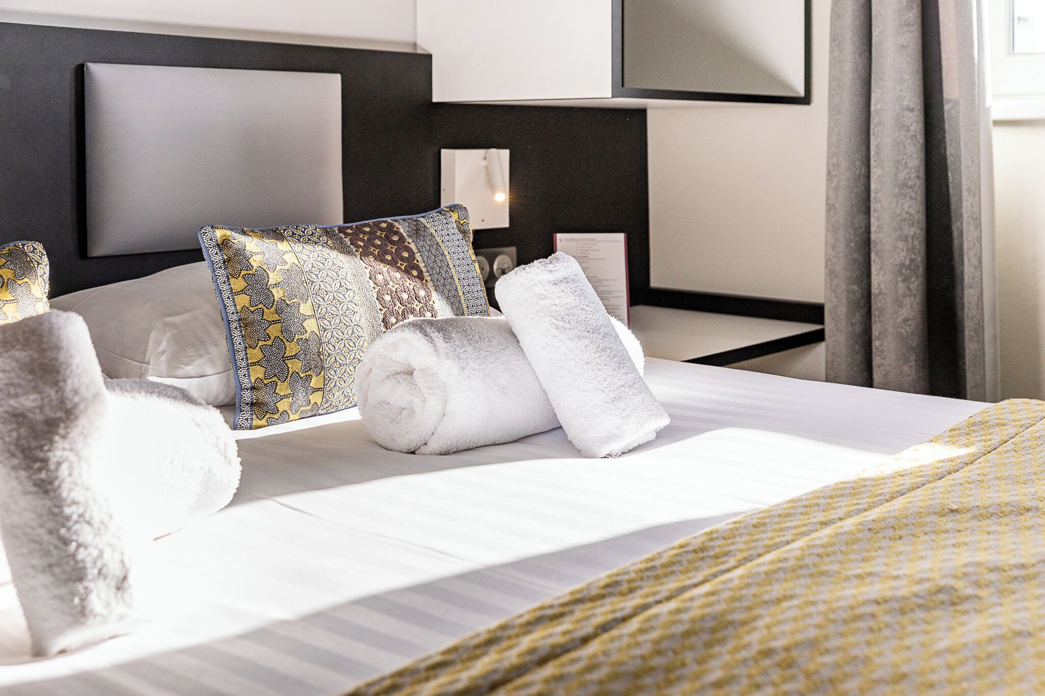 Een net opgemaakt bed met gestreepte kussens en opgerolde handdoeken, een nachtkastje met een lamp, en een modern hoofdbord.