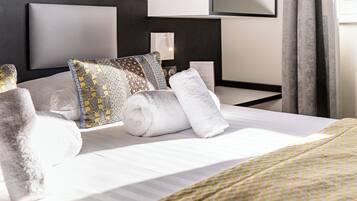 Een net opgemaakt bed met gestreepte kussens en opgerolde handdoeken, een nachtkastje met een lamp, en een modern hoofdbord.