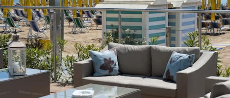 A beachside lounge with a sofa and a glass table, overlooking the beach with rows of sun loungers and umbrellas.