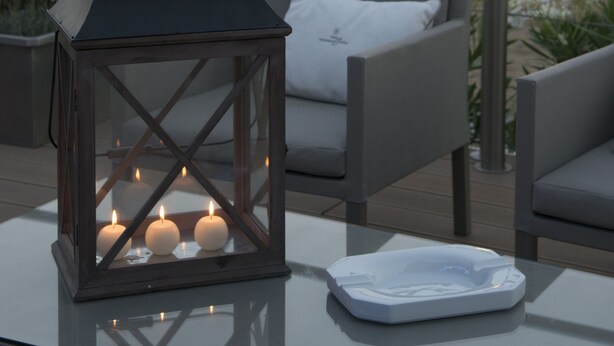 Uma mesa de vidro com um cinzeiro branco e um candeeiro aceso com velas.