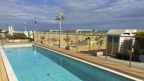 Piscina com águas cristalinas azuis, rodeada por um deck de madeira e uma área de praia arenosa com guarda-sóis e espreguiçadeiras.