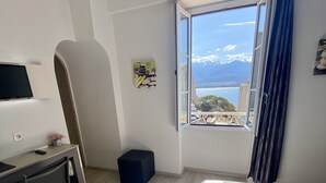 Ein Hotelzimmer mit Bett, Schreibtisch, Stuhl, Hocker, Mülleimer, einem Fenster mit Blick auf Meer und Berge sowie einem Bild an der Wand.