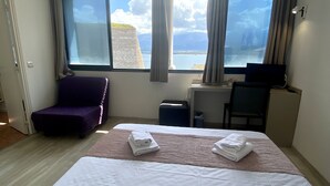 Ein Hotelzimmer mit großem Fenster und Meerblick, einem Bett mit weißer Bettwäsche, einem violetten Sessel und einem Schreibtisch mit Lampe.