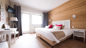A hotel room with a wooden wall, a bed with white bedding, a desk with a chair, and a TV mounted on the wall. - The Originals Boutique, Hotel Le Kastelberg (Andlau)
