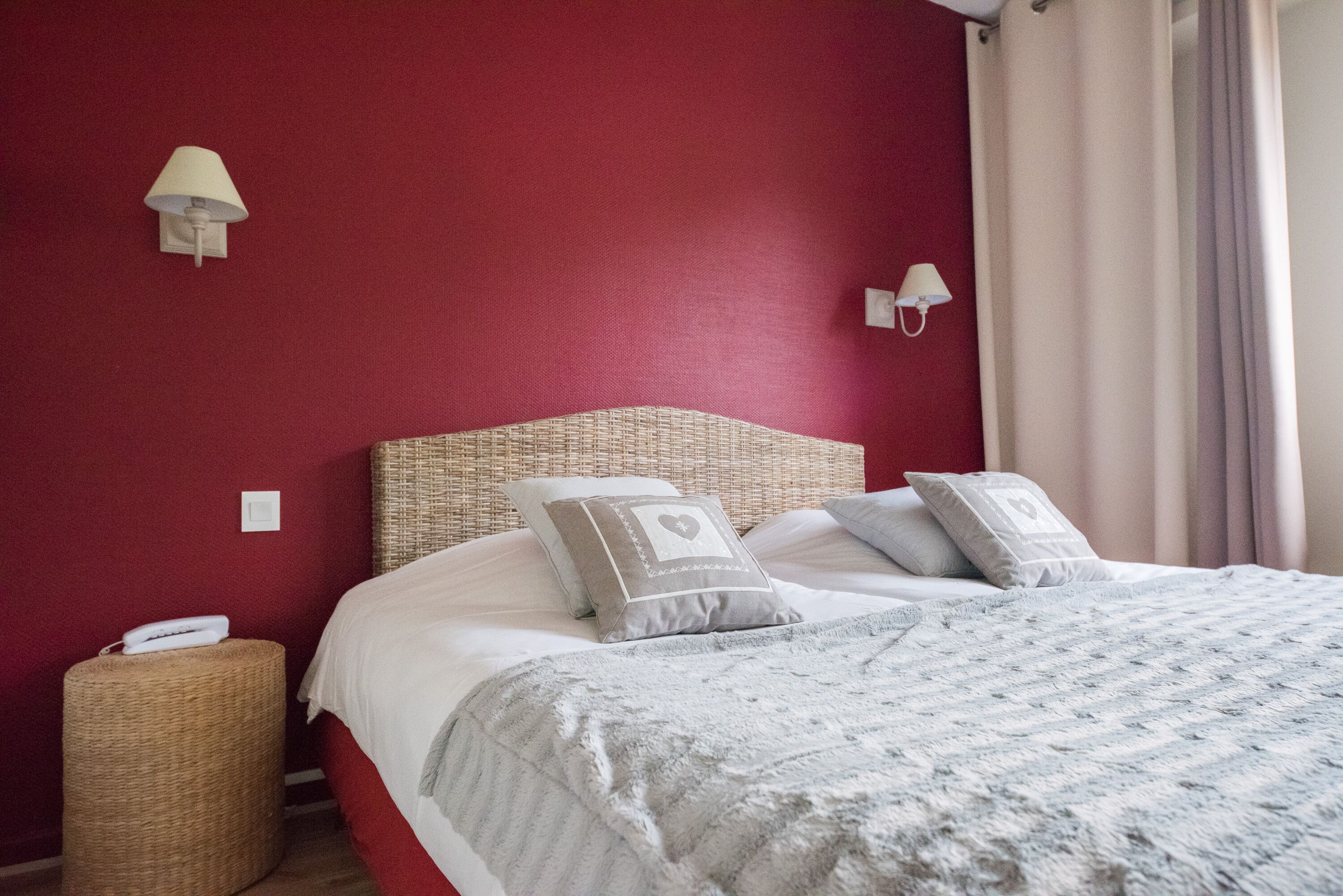 A neatly made bed with a grey blanket and two pillows, a wicker nightstand with a phone, and two wall-mounted lamps.
