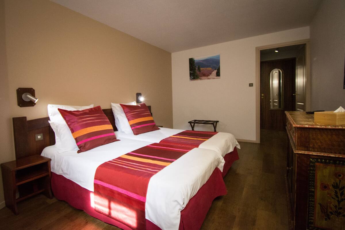Una habitació d'hotel amb un llit gran, una nit, una petita taula, un calaixera de fusta i una porta que condueix a una altra habitació.
