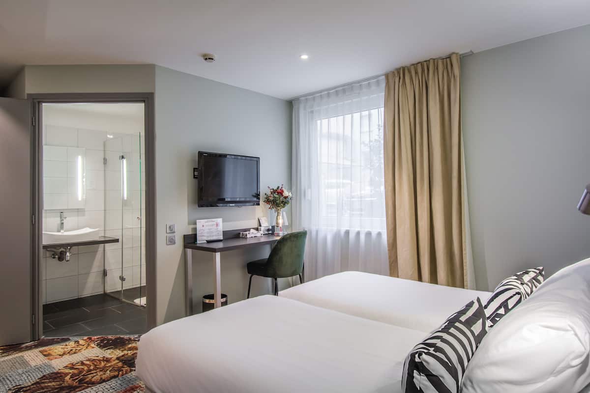 A hotel room with a bed, a desk with a computer, a television, and a bathroom visible through an open door.