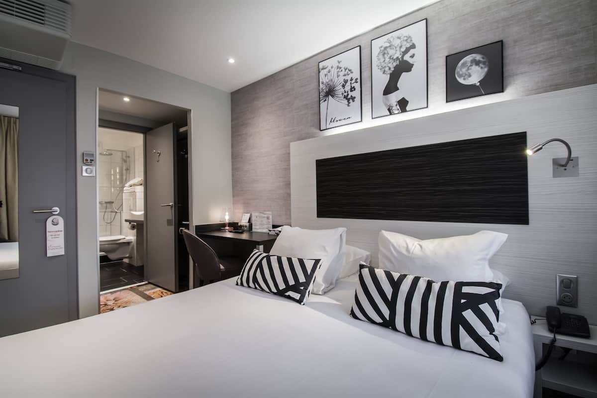 A modern hotel room with a bed, bedside table, and a desk with a chair. There are framed pictures on the wall and a bathroom visible through an open door.