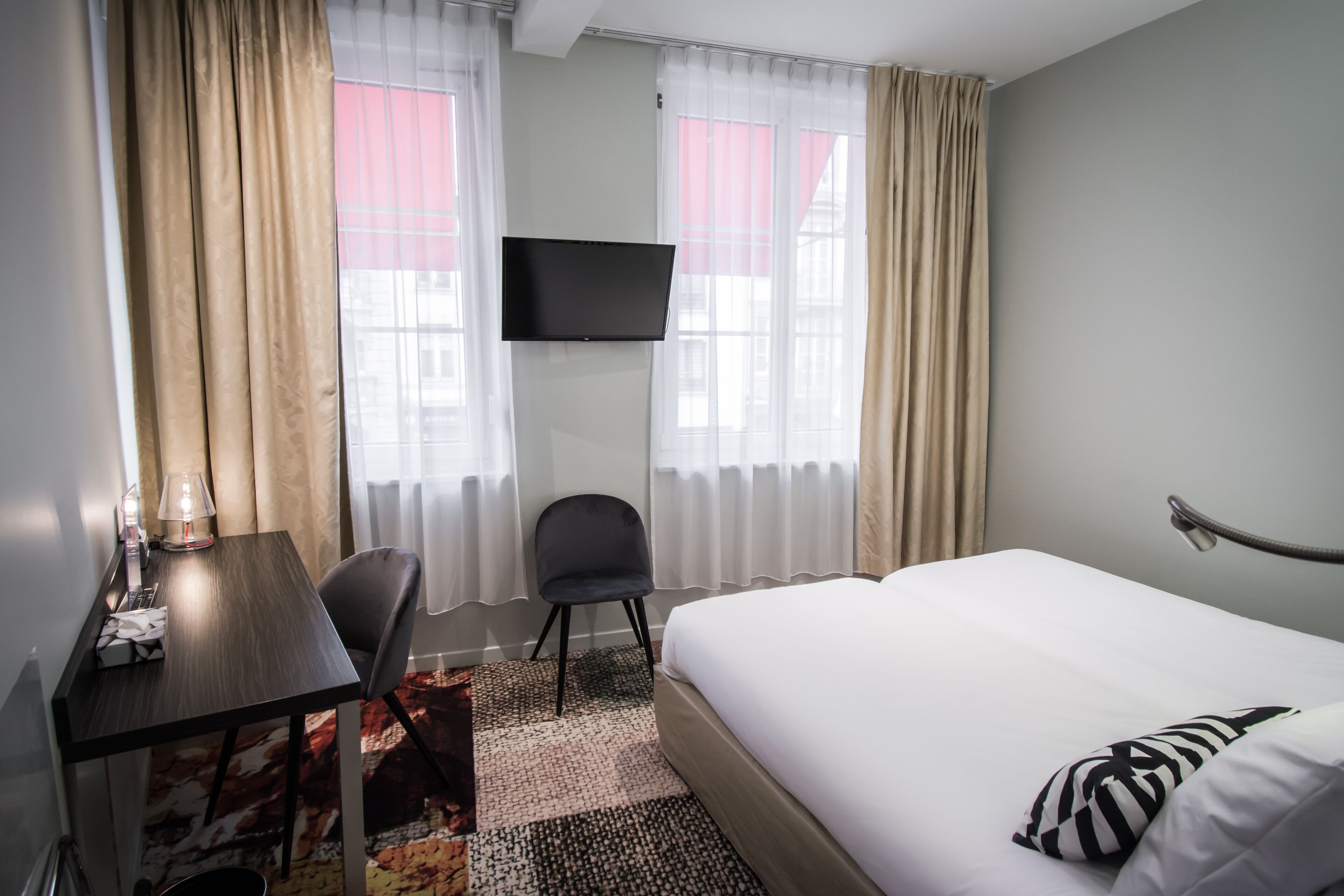 A hotel room with a bed, a desk with a chair, a flat-screen TV mounted on the wall, and a window with curtains.