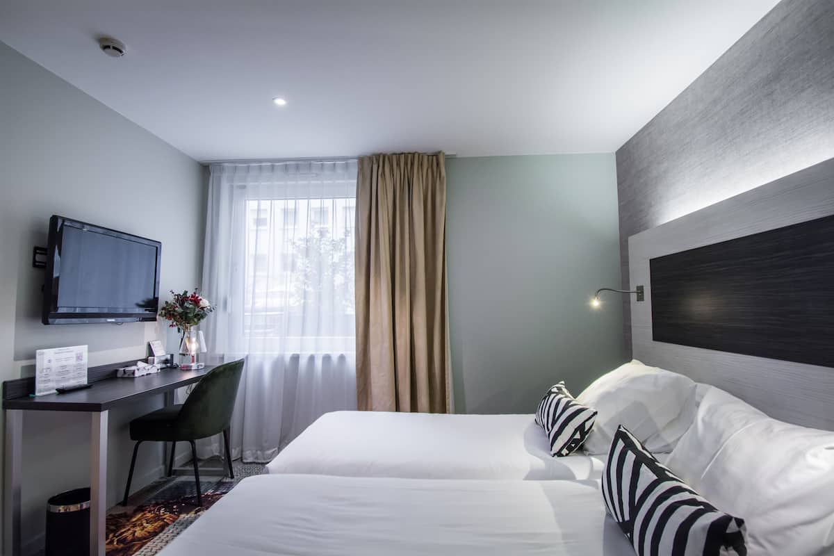 A hotel room with a bed, a desk, a chair, a TV, and a window with curtains.