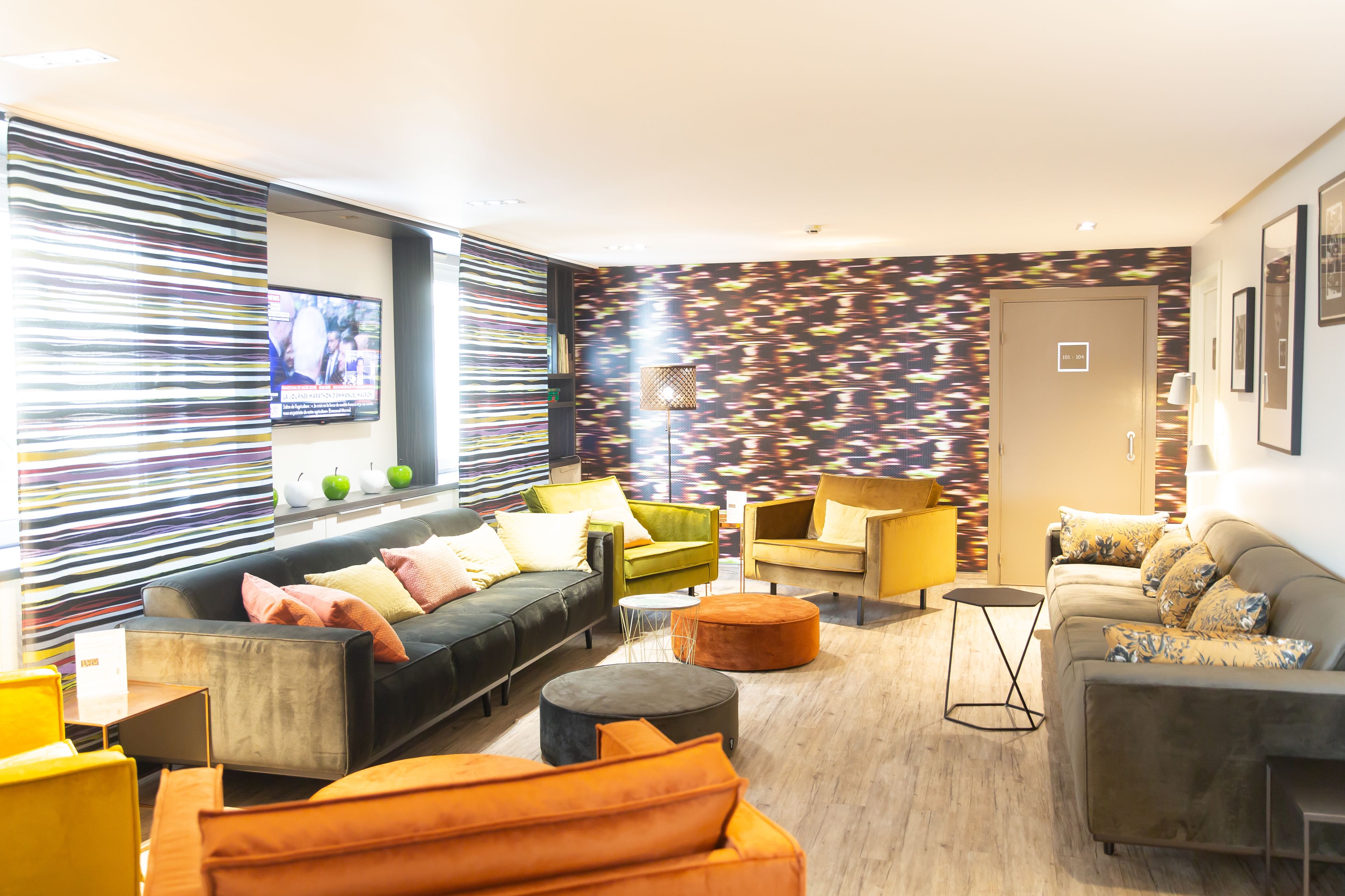 Ein modernes Wohnzimmer mit einer Couch, Sesseln und Poufs. Ein Fernseher ist an der Wand montiert und es gibt eine Wand mit Muster hinter der Couch.