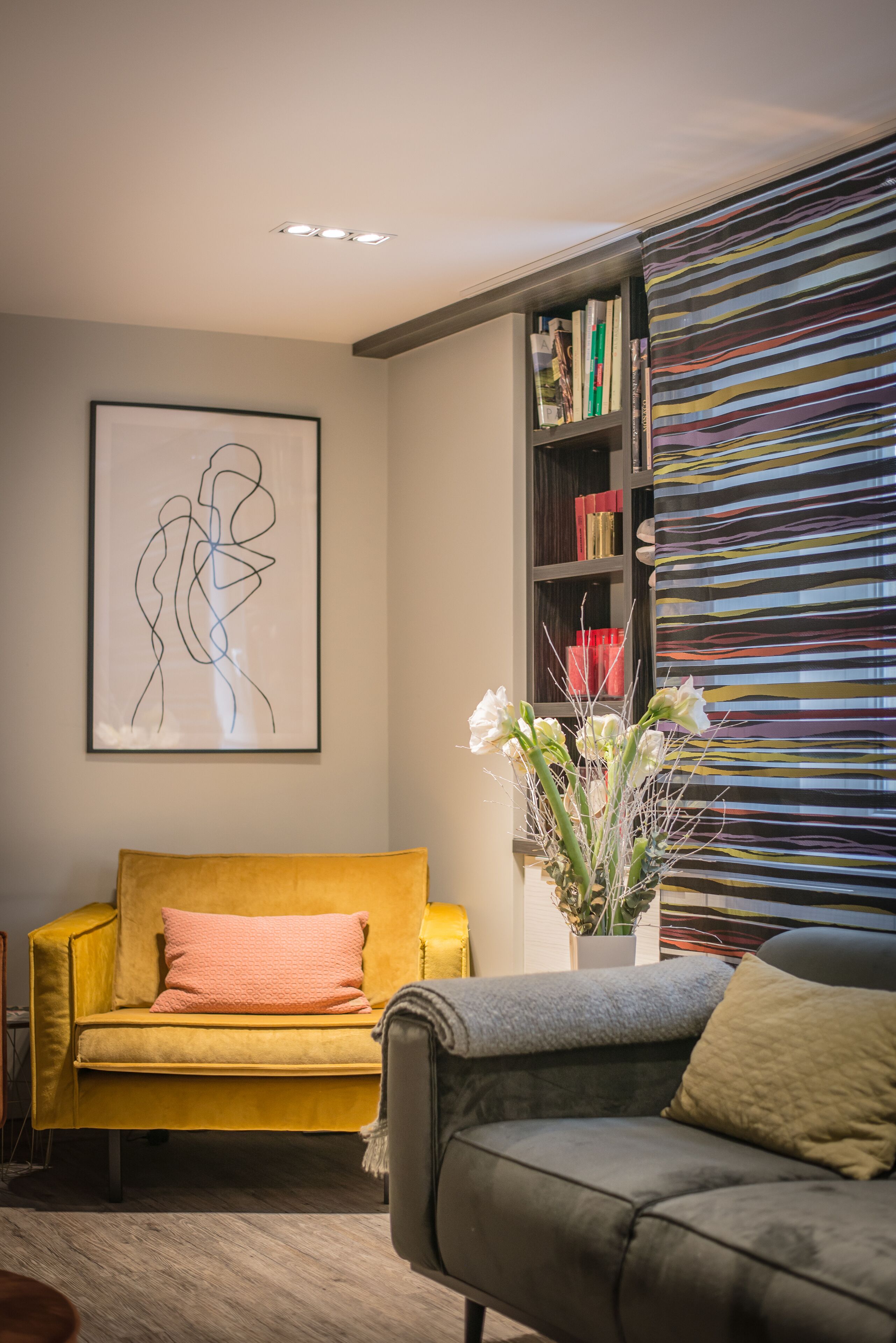 Una sala de estar con un sofá amarillo, un sofá gris, una estantería y un cuadro abstracto enmarcado.