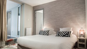 A modern bedroom with a large bed, a nightstand with a lamp, and a sliding glass door leading to a balcony. - Couvent du Franciscain (Strasbourg)