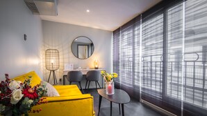 A modern hotel room with a yellow sofa, a round mirror, a small table with a vase of flowers, and a large window with blinds. - Couvent du Franciscain (Strasbourg)