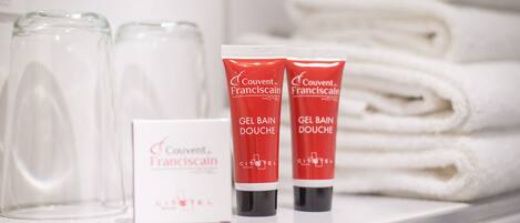 A hotel shelf with toiletries including two red tubes labeled 'Couvent Franciscain Gel Bain Douche' and a white box with the same branding.