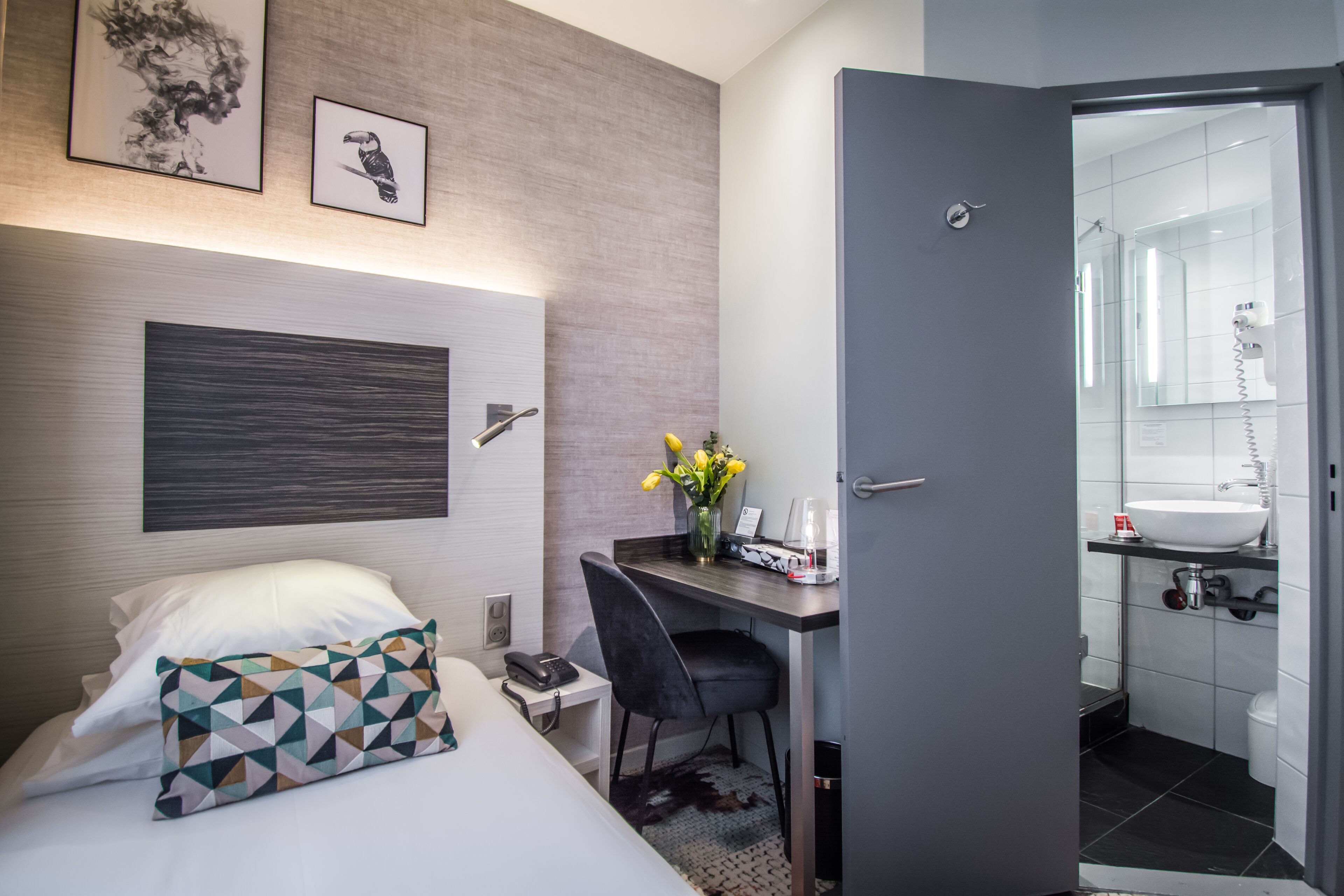 Habitación de hotel con una cama, un escritorio, una silla, un jarrón con flores, un teléfono y una puerta que da a un baño.