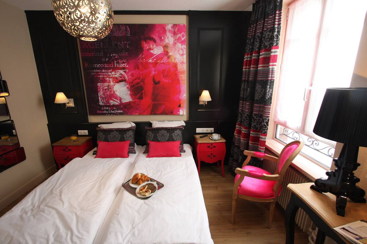 A hotel room with a large bed, two bedside tables with lamps, a chair, a desk with a lamp, and a window with curtains.