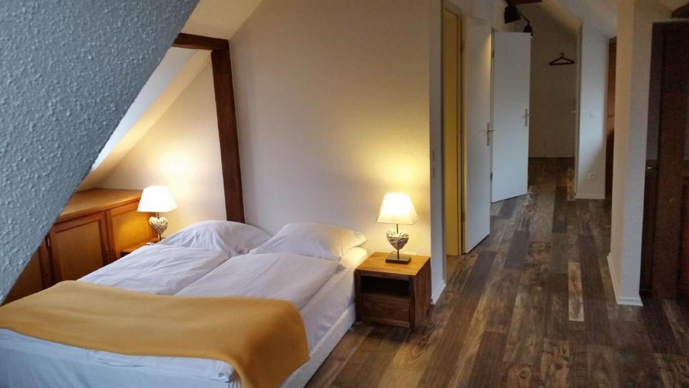 A bedroom with a sloped ceiling, two bedside lamps, a bed with white linens, and a wooden floor.