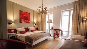 Habitación de hotel con una cama grande, dos mesitas de noche, un escritorio y una silla. Se aprecia una araña, una ventana con cortinas y una obra de arte roja en la pared.