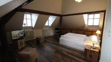 Une chambre avec un plafond de style industriel, des poutres en bois, un lit avec du linge de lit blanc, une table de chevet avec une lampe, un téléviseur posé sur un meuble en bois et une fenêtre donnant sur l’extérieur.