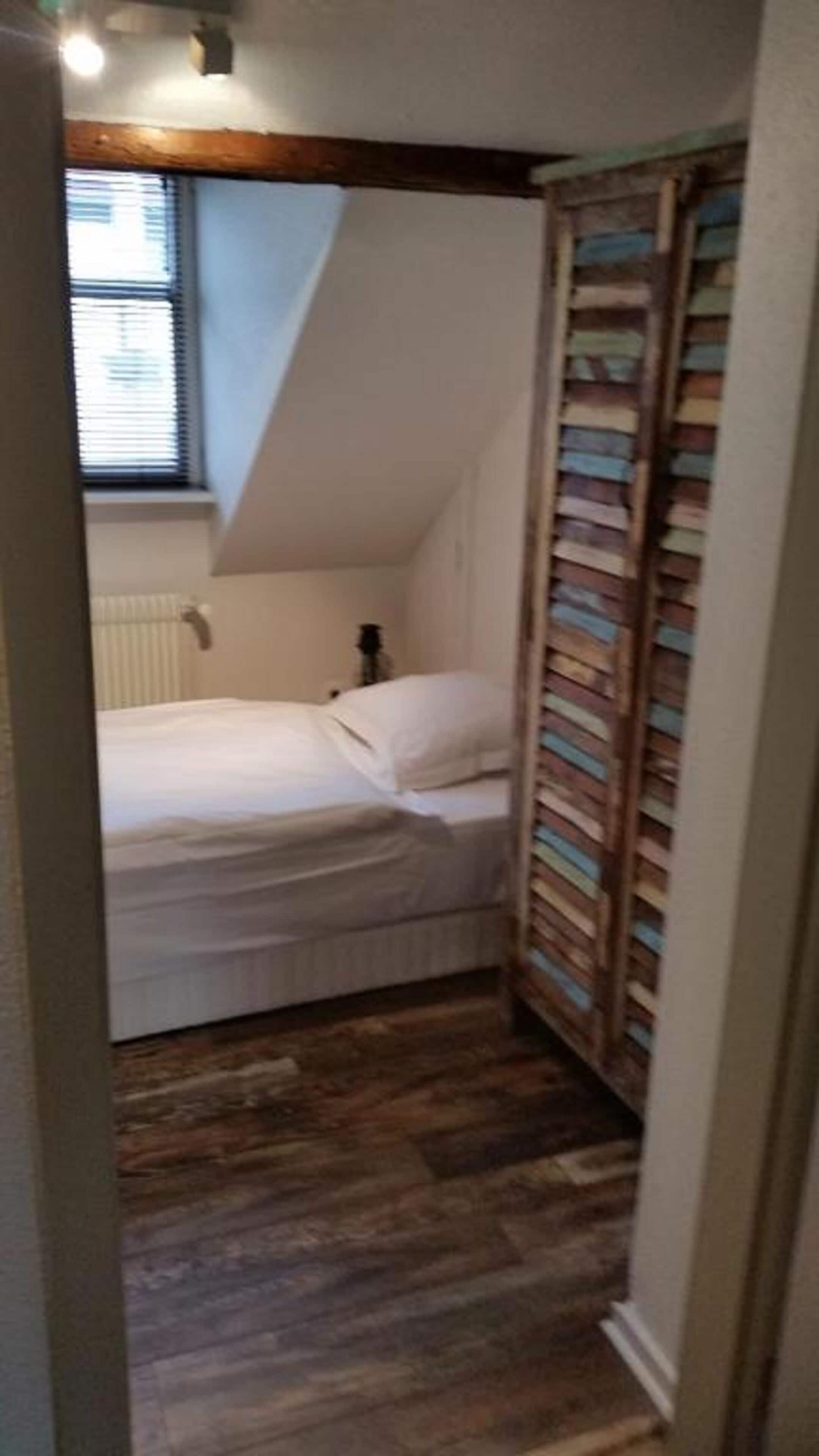 A small, narrow room with a sloped ceiling, a bed, a radiator, and a window with blinds.