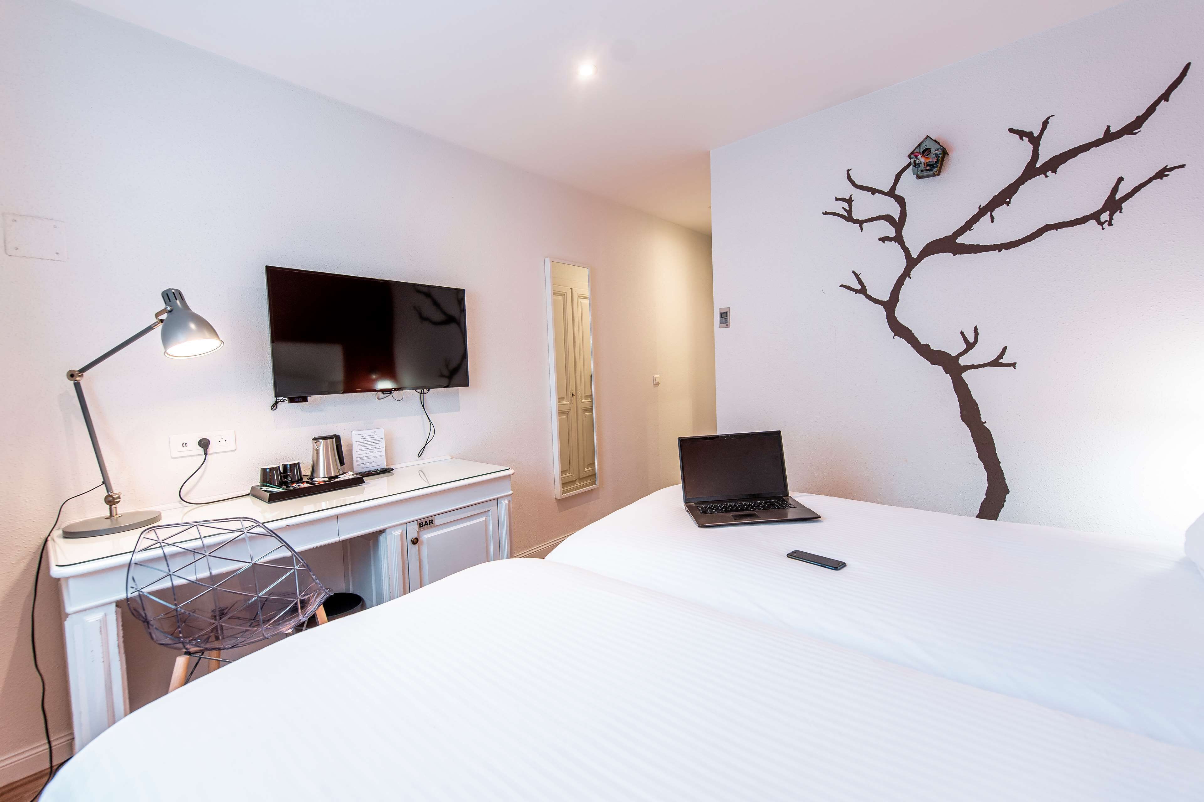 A hotel room with a bed, a desk with a laptop, a TV, and a wall-mounted artwork of a tree.