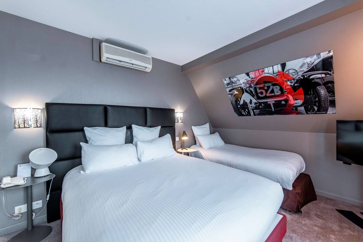A hotel room with two beds, a mounted TV, a wall-mounted air conditioner, and a motorcycle-themed artwork.