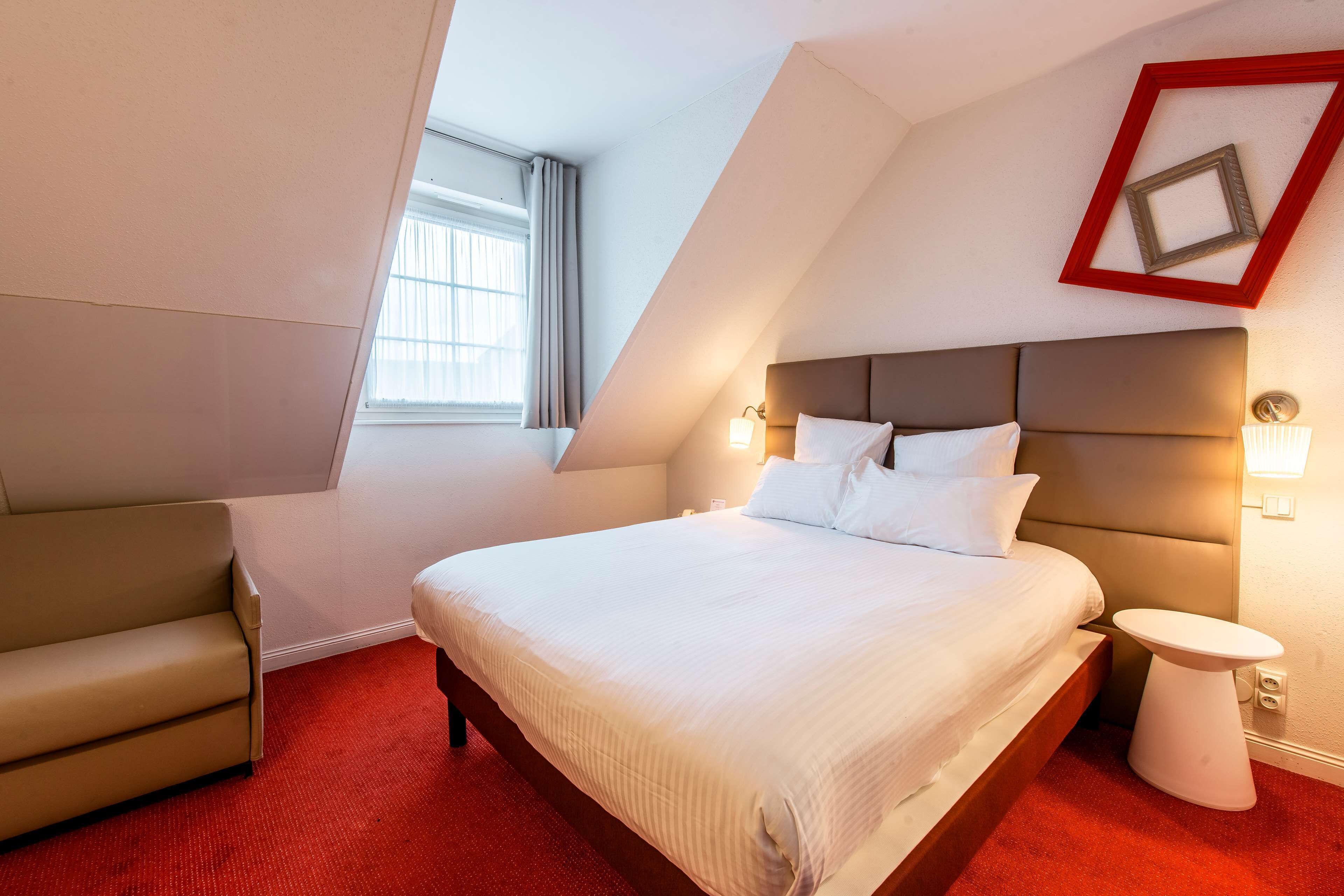Una habitación de hotel con una cama grande, un sillón pequeño, una mesita de noche y una ventana con cortinas.