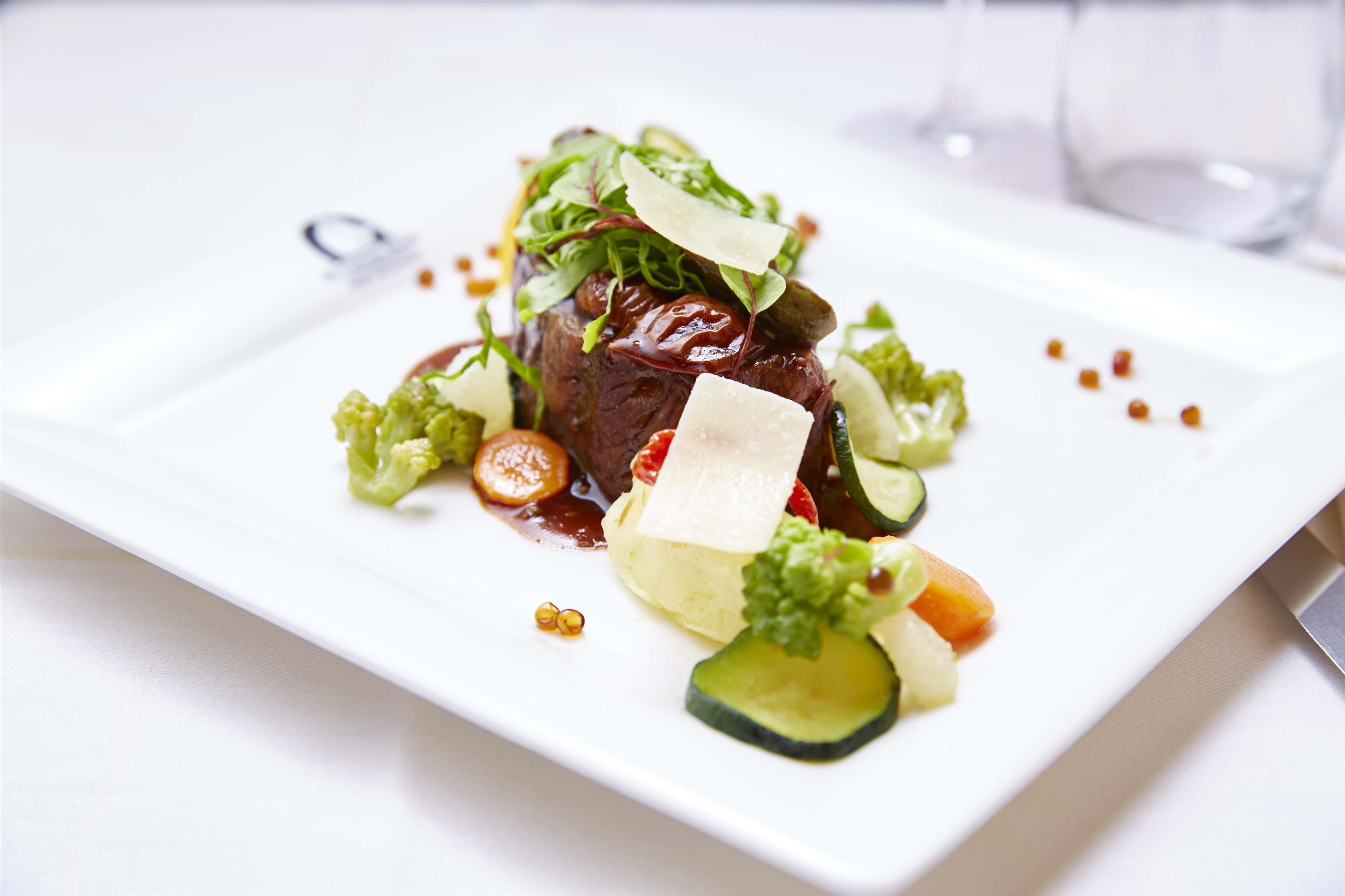 A white plate with a steak topped with greens, cheese, and vegetables.