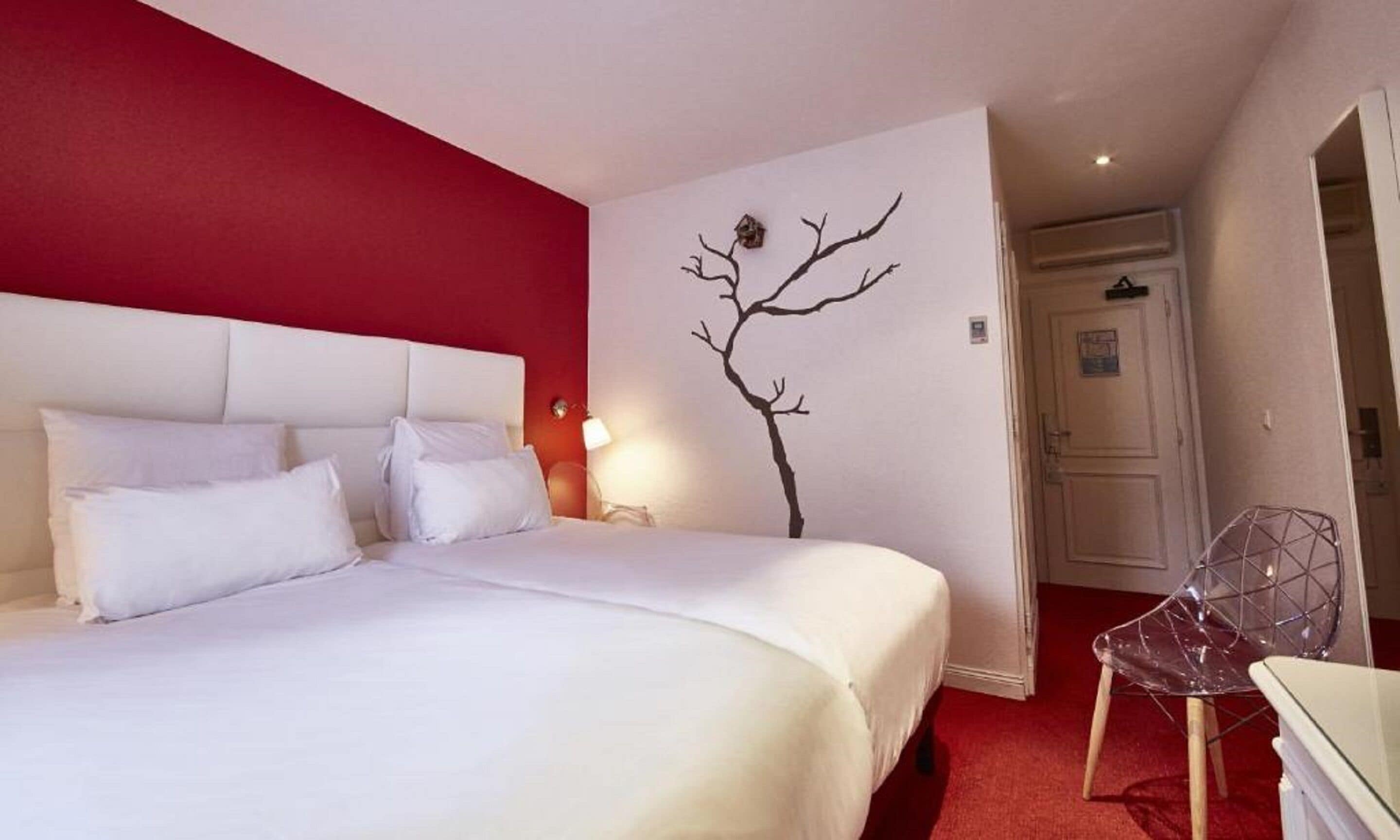 Une chambre d’hôtel avec un grand lit, un mur d’accent rouge, une décoration murale représentant un arbre et une chaise transparente.
