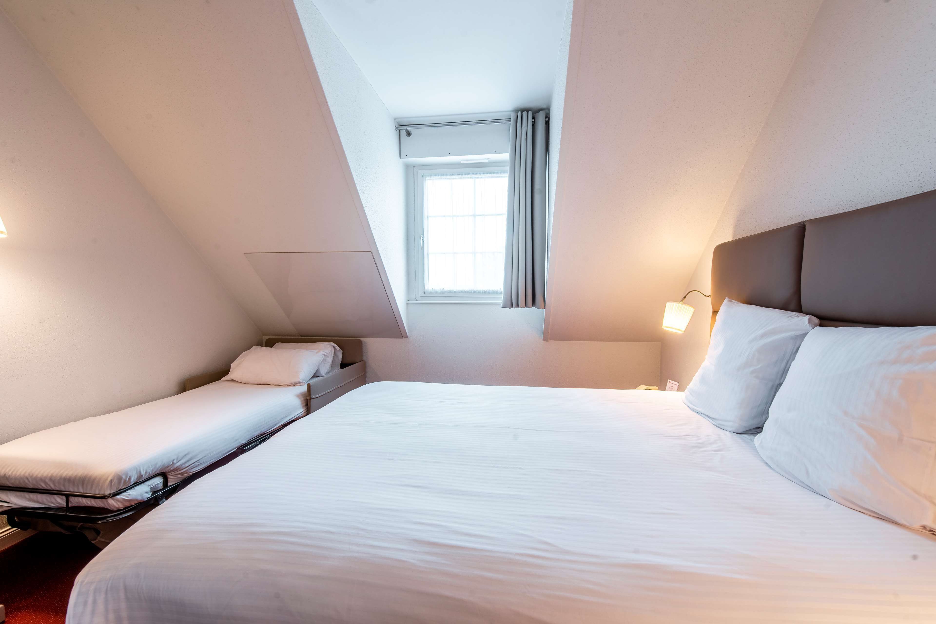 Ein Hotelzimmer mit einem großen Bett, einem kleineren Bett, einem Fenster und einem Nachttisch mit einer Lampe.