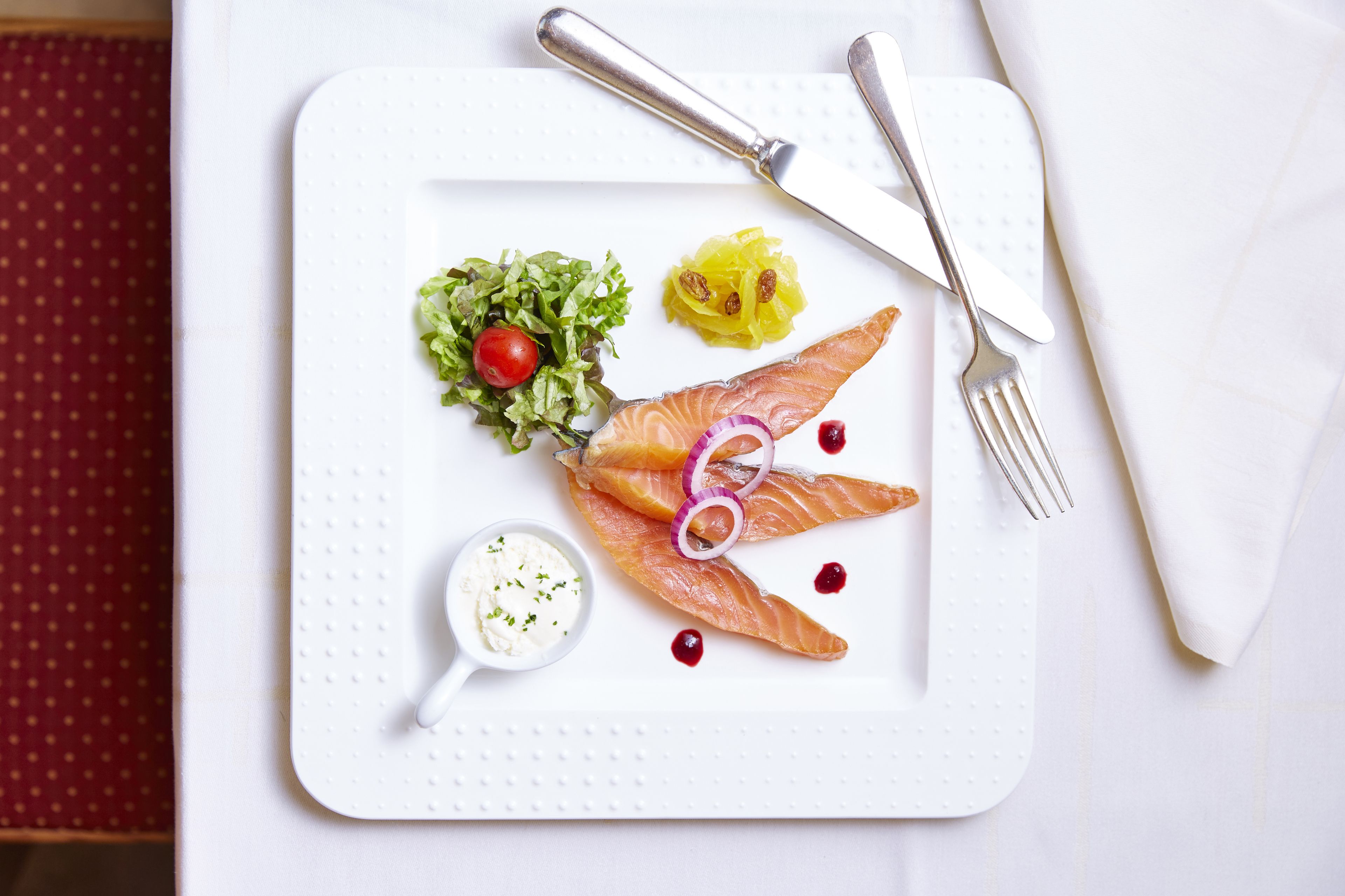 A white plate with salmon, a salad, and a small bowl of sauce, accompanied by a fork and knife on a white napkin.