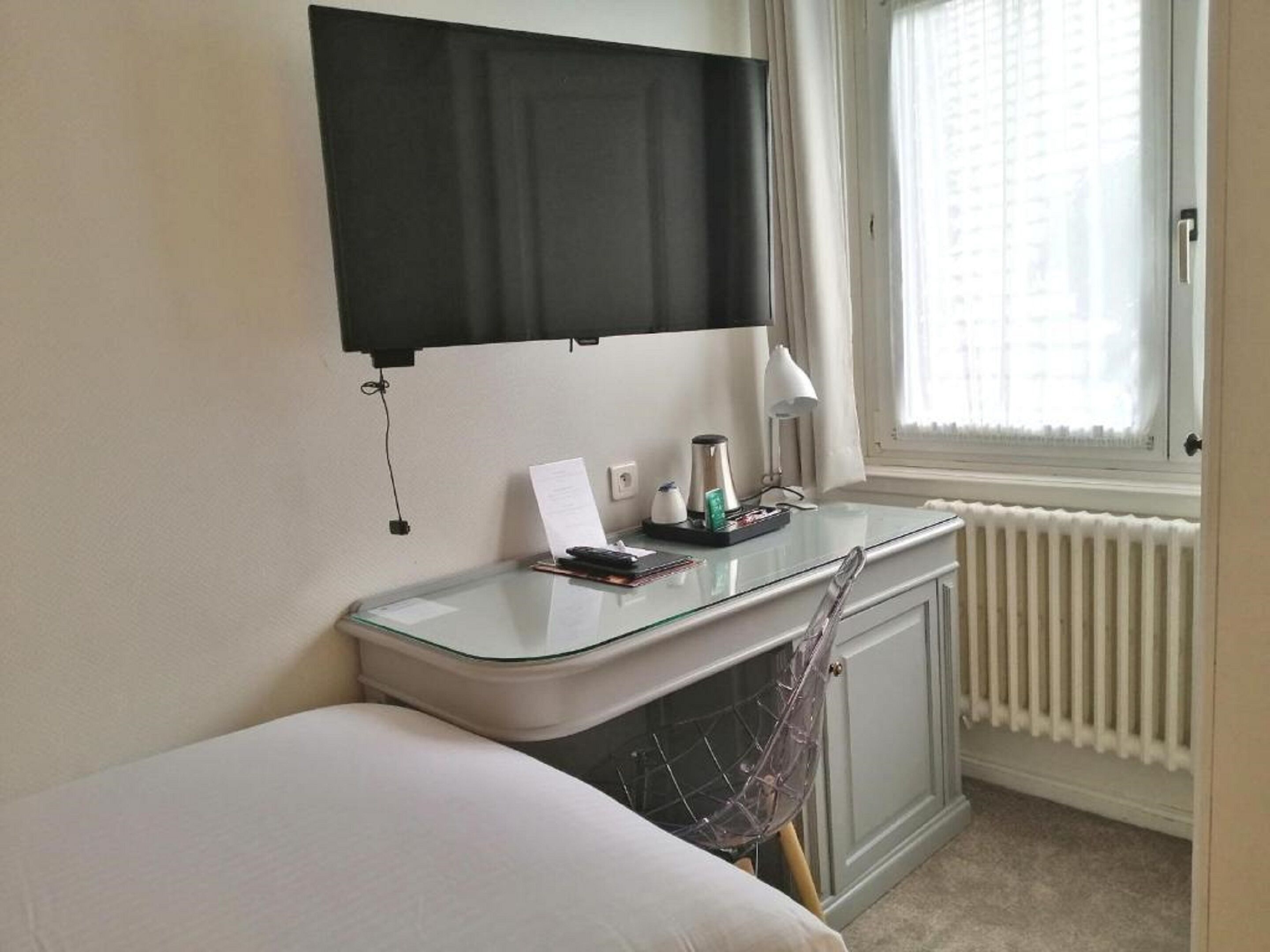 A compact hotel room with a glass desk, a bed, a radiator, and a window with curtains.