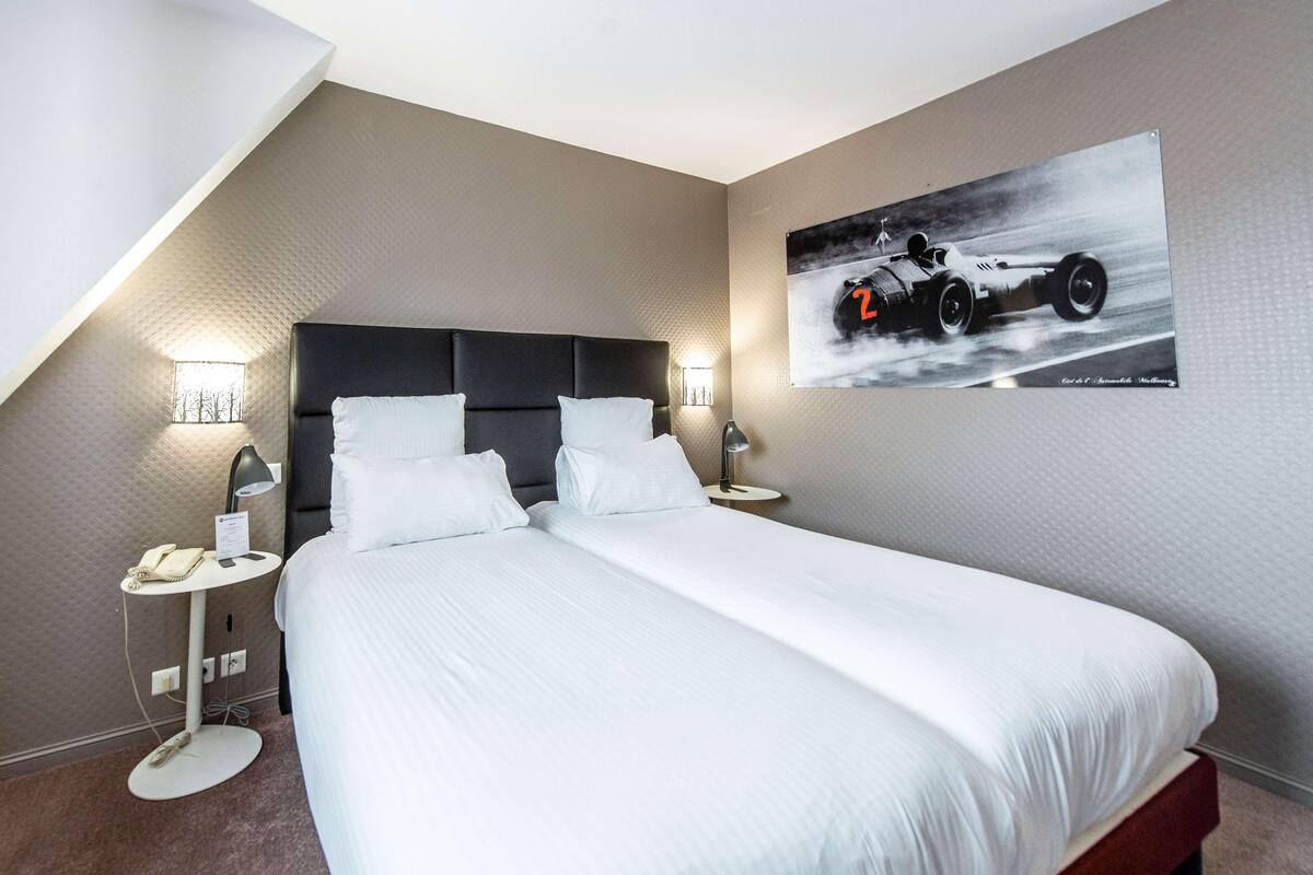 A modern hotel room with a large bed, two bedside tables with lamps, a small round table with a telephone, and a wall-mounted picture of a racing car.