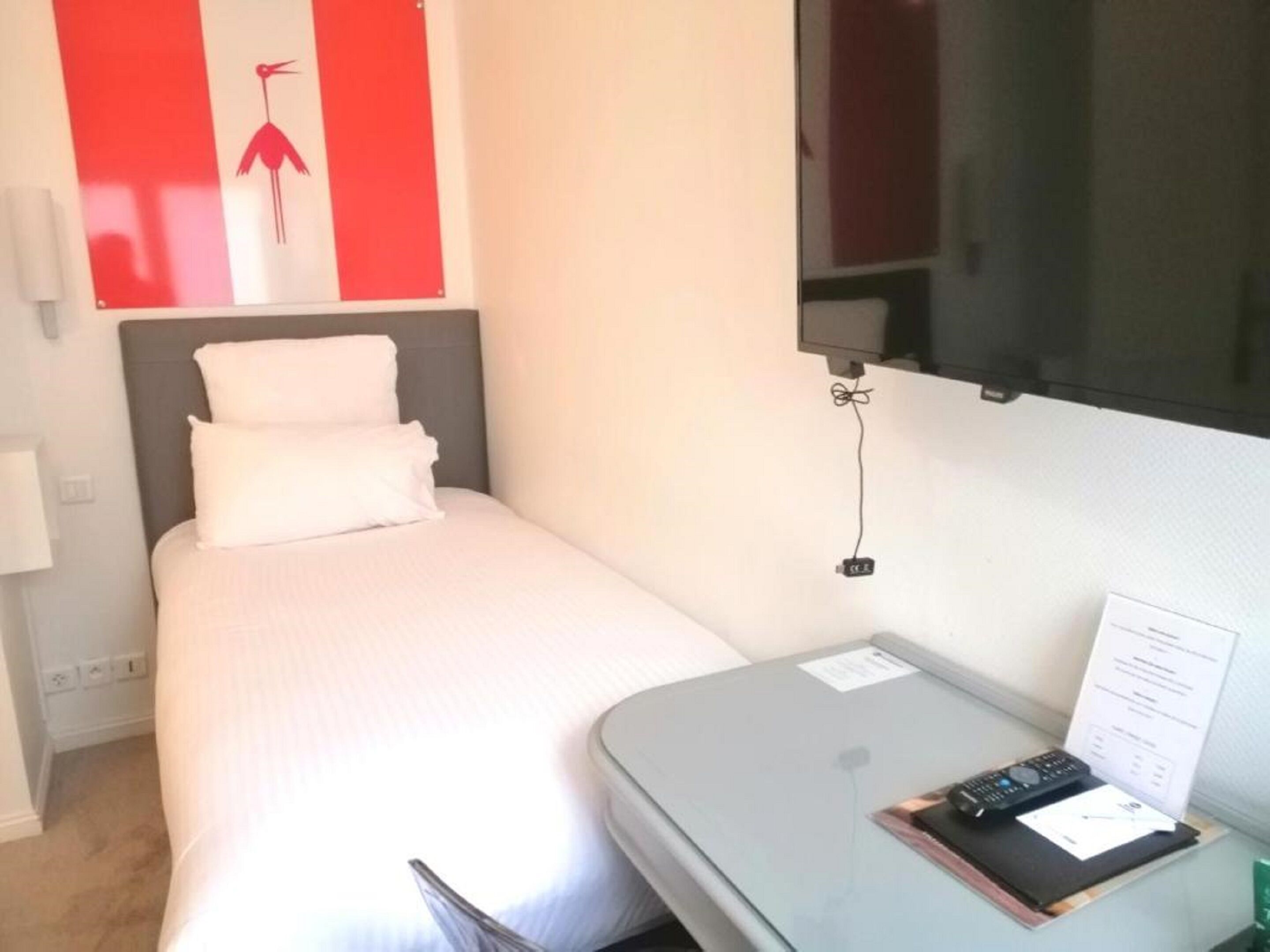A small, neatly made bed with white linens in a room with a grey headboard. There is a small table with a remote control and a piece of paper on it.