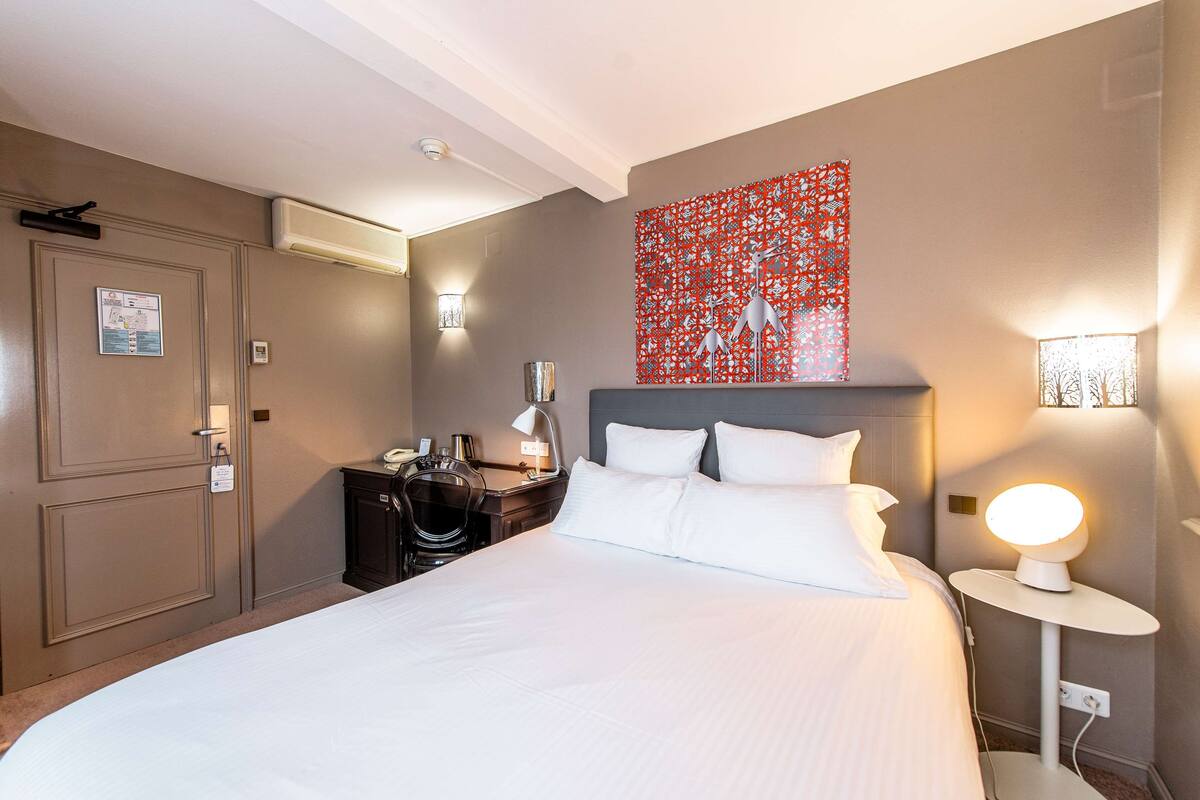 A hotel room with a large bed, a desk, a chair, a wall-mounted lamp, and an air conditioning unit.