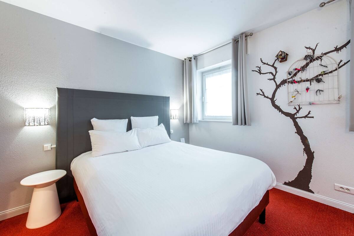 A hotel room with a large bed, a small bedside table, and a wall mural of a tree with birds.