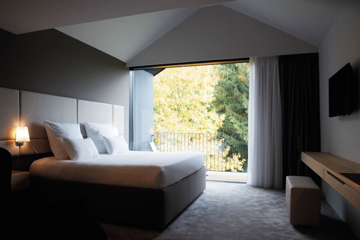 A modern bedroom with a large bed, a bedside table, a desk, and a view of trees through a window.