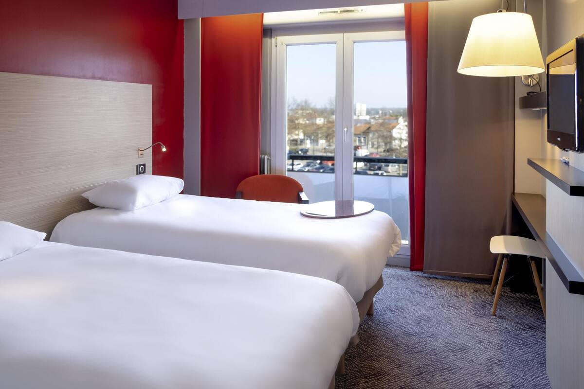 Habitación de hotel con dos camas, un escritorio, una silla, un televisor y una ventana con vista a la ciudad.