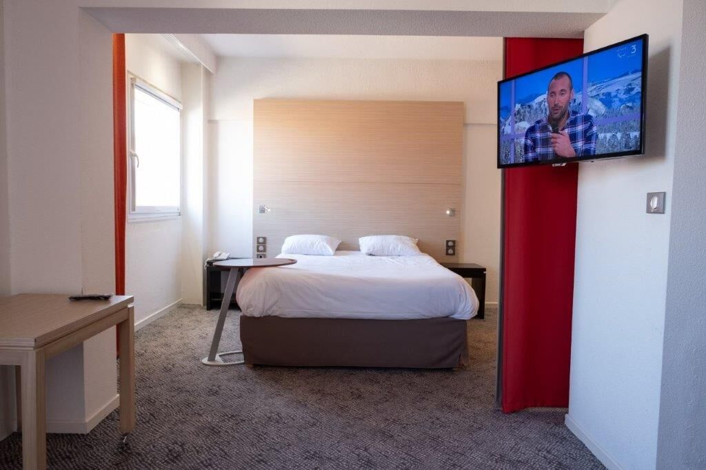 Una habitación de hotel con cama, televisor montado en la pared, escritorio y ventana.