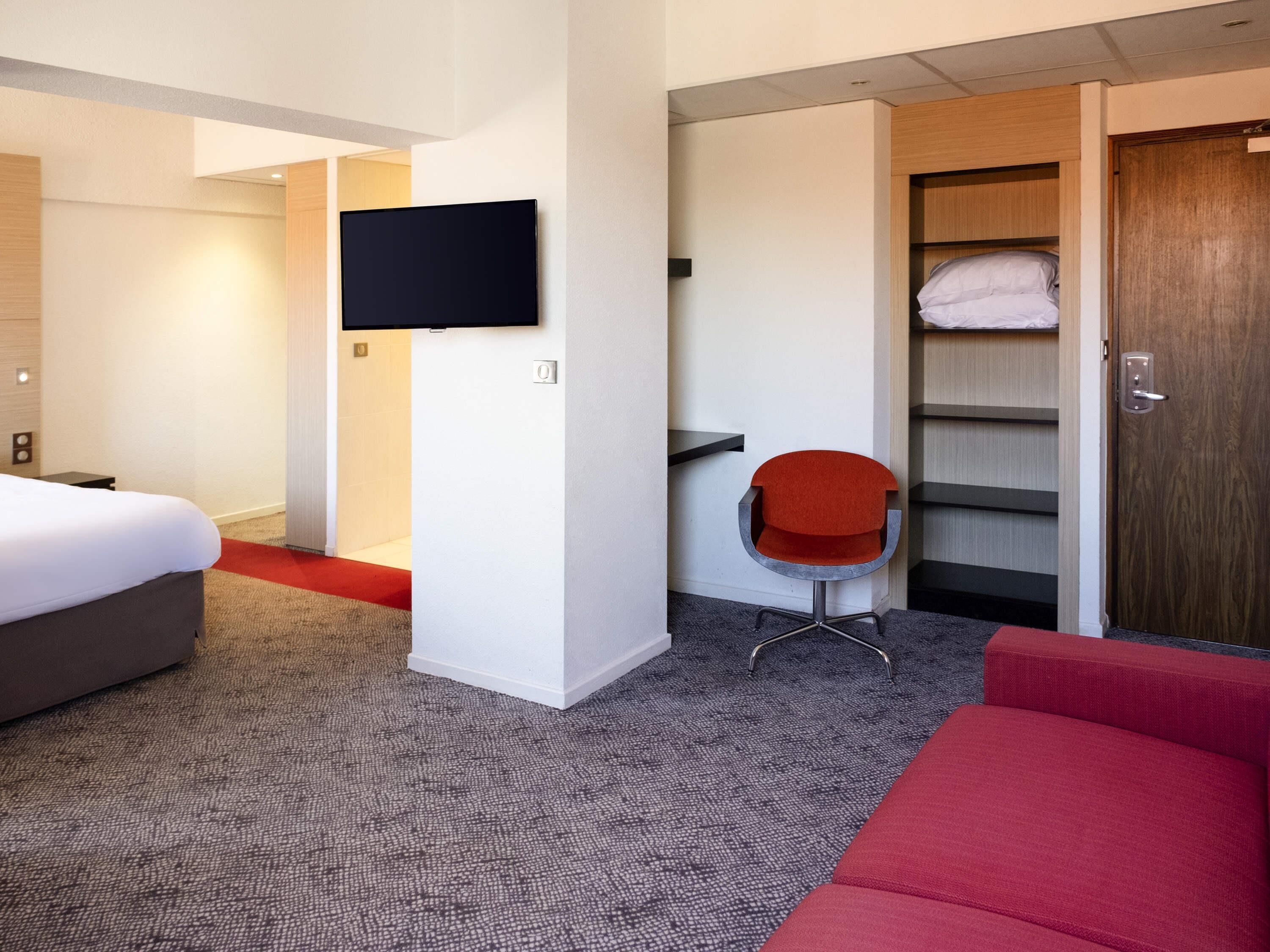 Et hotelværelse med en seng, et fjernsyn ophængt på væggen, en rød stol, et garderobeskab og en rød sofa.